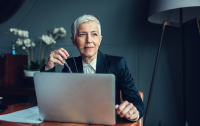 Portrait of mature businesswoman, sitting in her office and thinking. Holding eyeglasses in her mouth. In front of her on the desk is laptop.