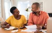 Cheerful mature couple sitting and managing expenses at home. Happy african man and woman paying bills together and managing budget. Black smiling couple checking accountancy and bills while looking at each other. (Cheerful mature couple sitting and m